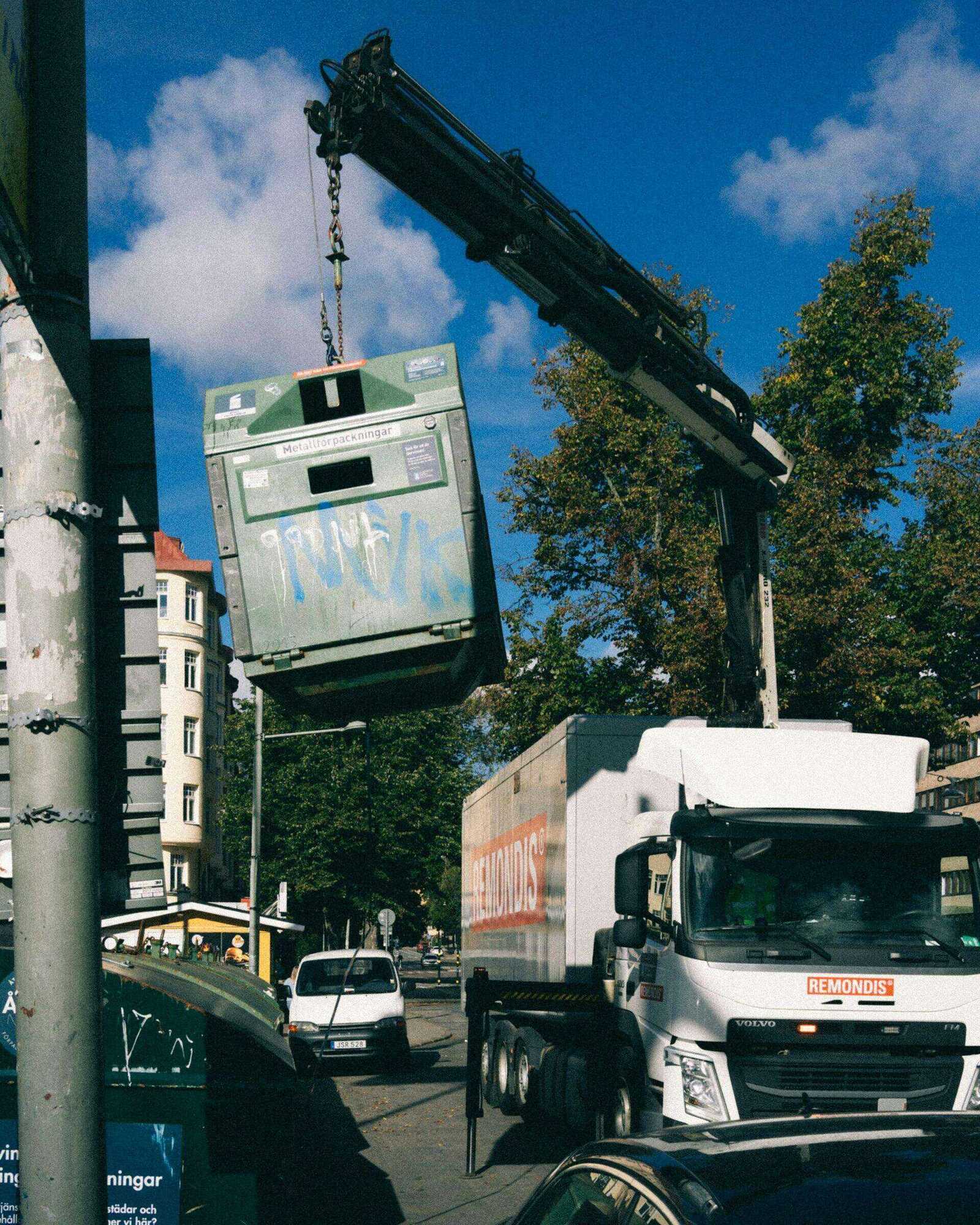 Crane truck lifting a large recycling bin onto a waste collection vehicle on a city street